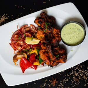 Tandoori kemencében enyhén kormosra sült csirkehús, ecetesd lilahagyma, zöldségkeverék, és egy kis tálka mentás csatni egy fehér tányéron tálalva. Mindez körbeszórva egy kis köménnyel, csillagánizzsal, és szárított chilivel.